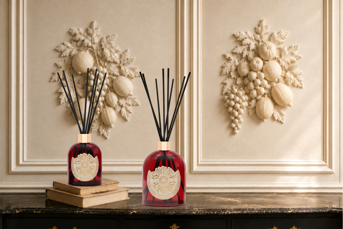 Deux bouquets parfumés Saint Songe rouges et or, avec leurs bâtonnets noirs, posés sur une console devant des ornements muraux et des livres anciens.